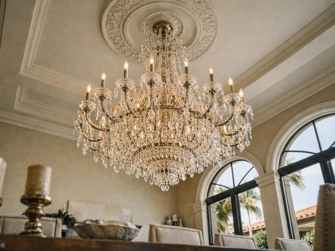 A crystal chandelier hangs from an ornate ceiling above a bright room with arched windows and dining chairs