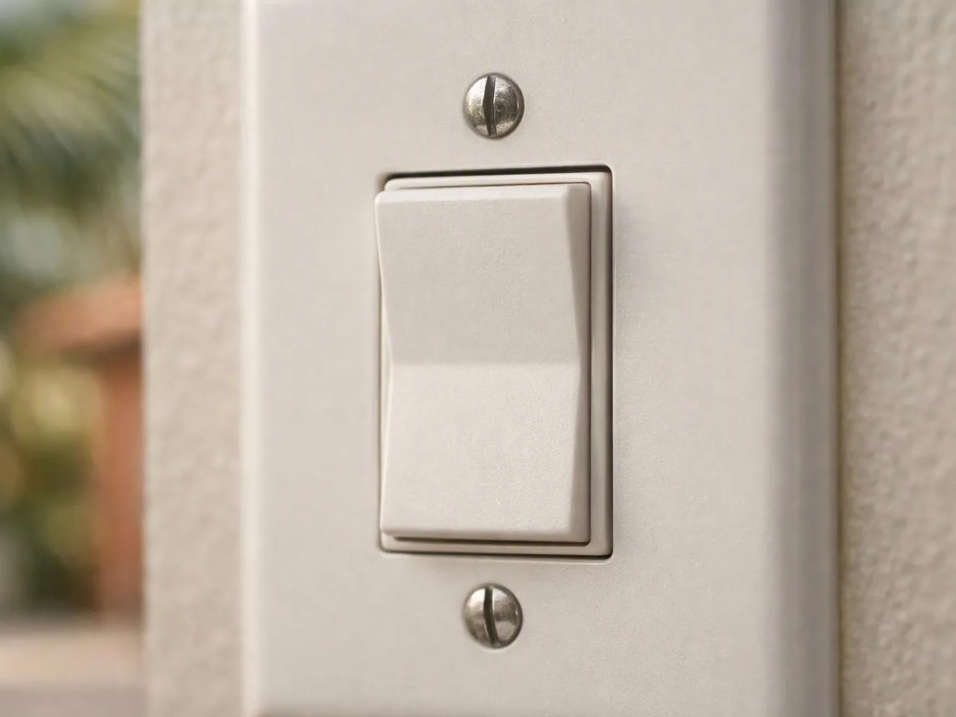 A white wall light switch sits on a beige wall with blurred background shapes to the left